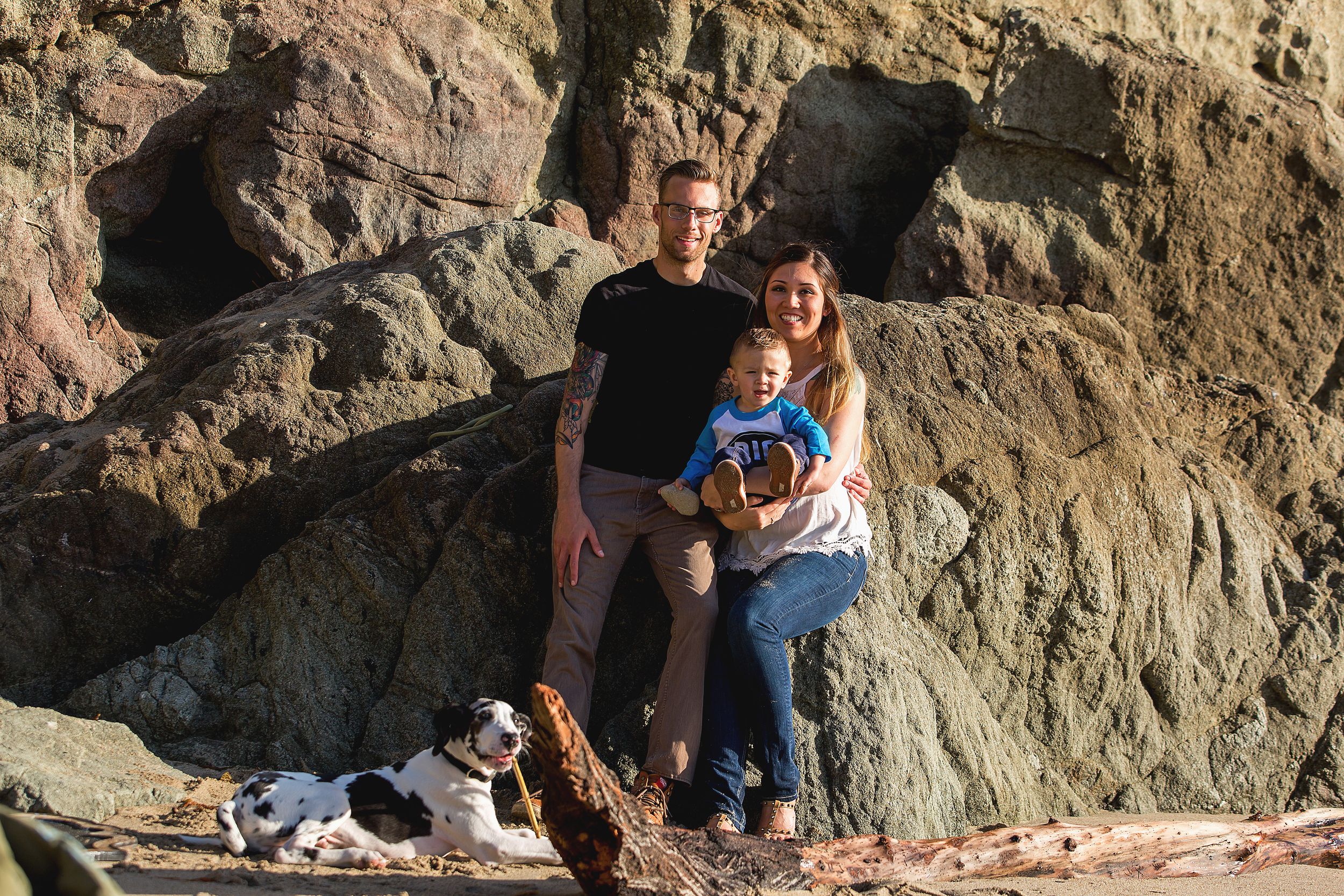 A couple poses with their dog against large coastal rocks at sunset.