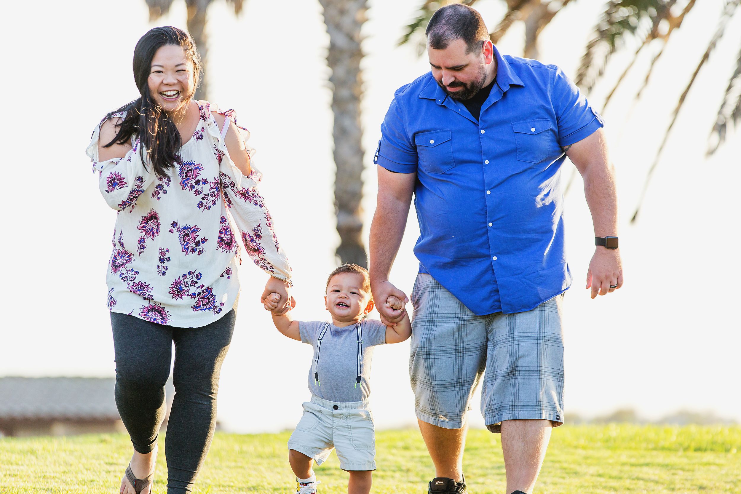 A family walks together holding hands on a grassy path with palm trees.
