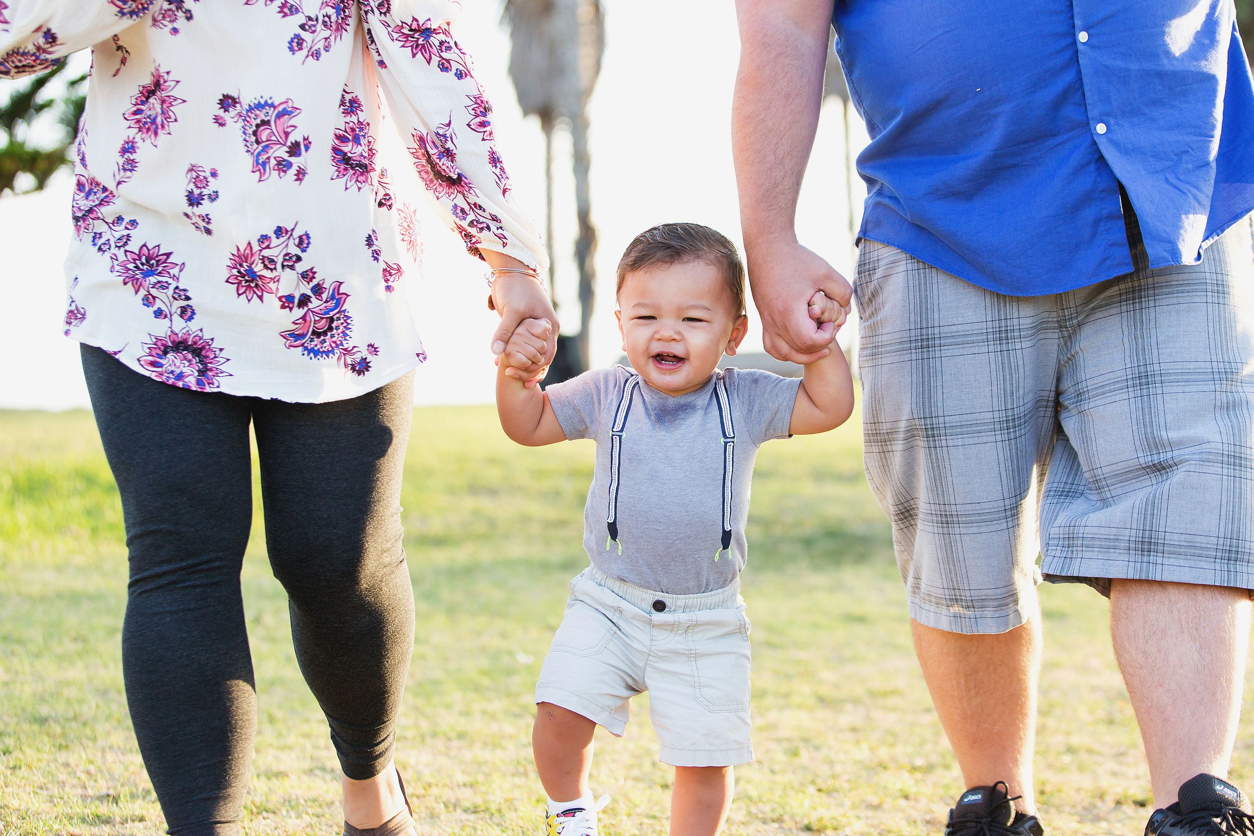 A toddler learns to walk while holding hands with adults on a sunny day.