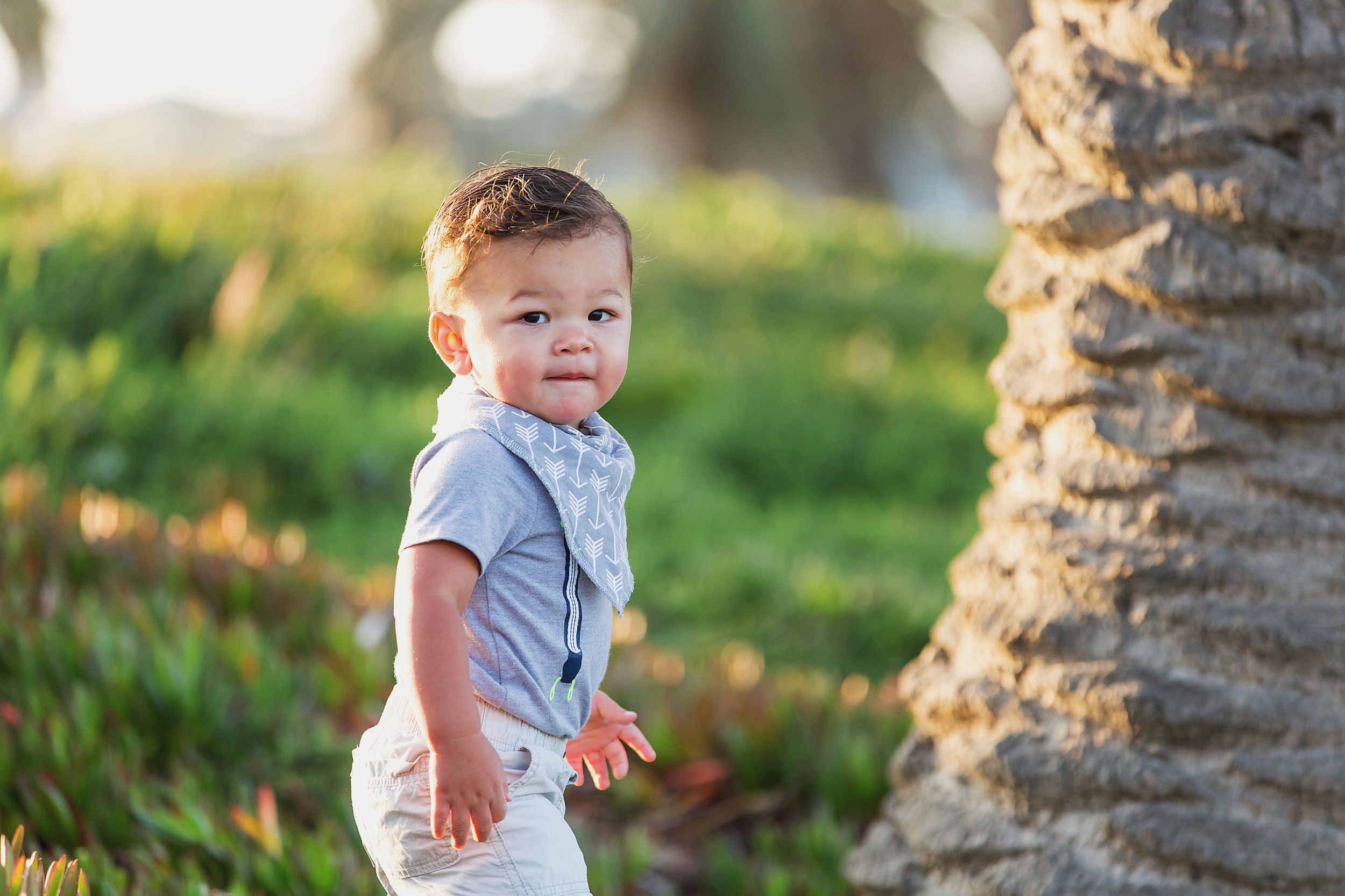A cheerful toddler in light colored clothing stands next to a palm tree trunk.