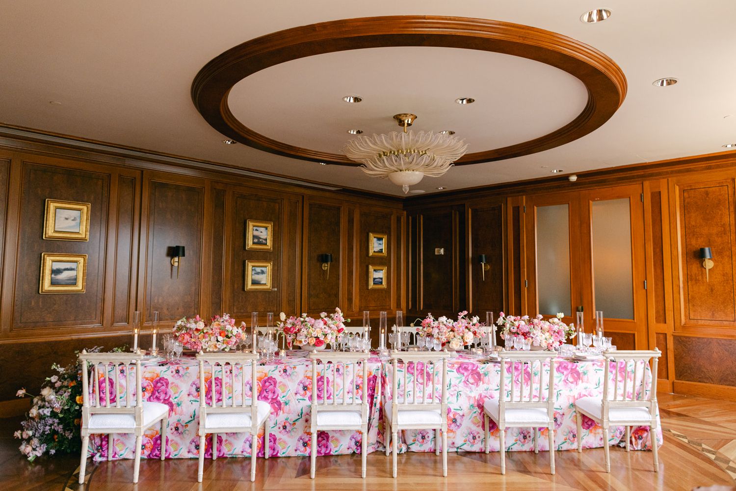 An elegant banquet room with wood-paneled walls features long tables decorated with pink floral arrangements and white chairs.