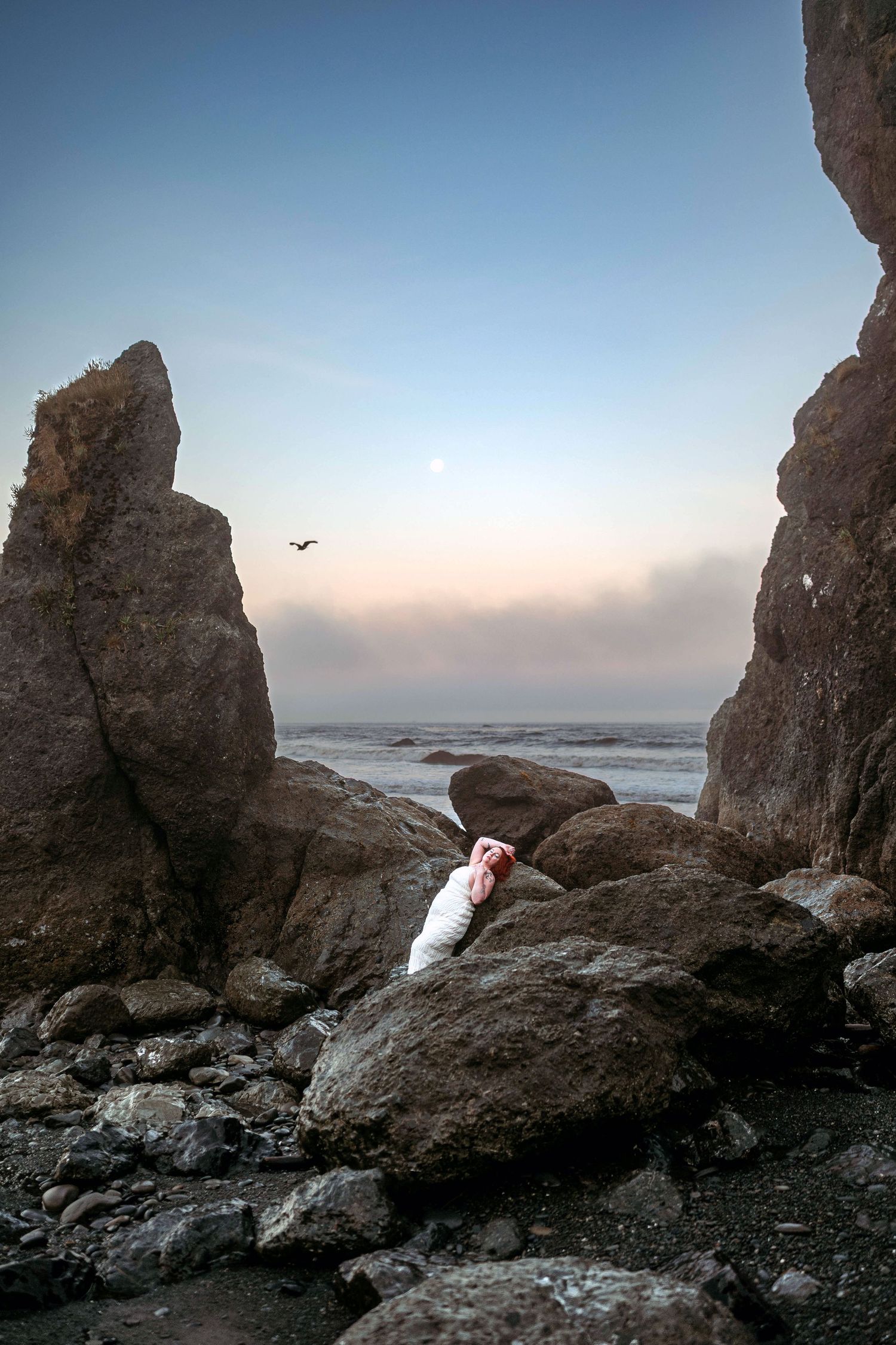 A woman in a white dress photographed between coastal rock formations at sunrise. Fantasy photoshoot.