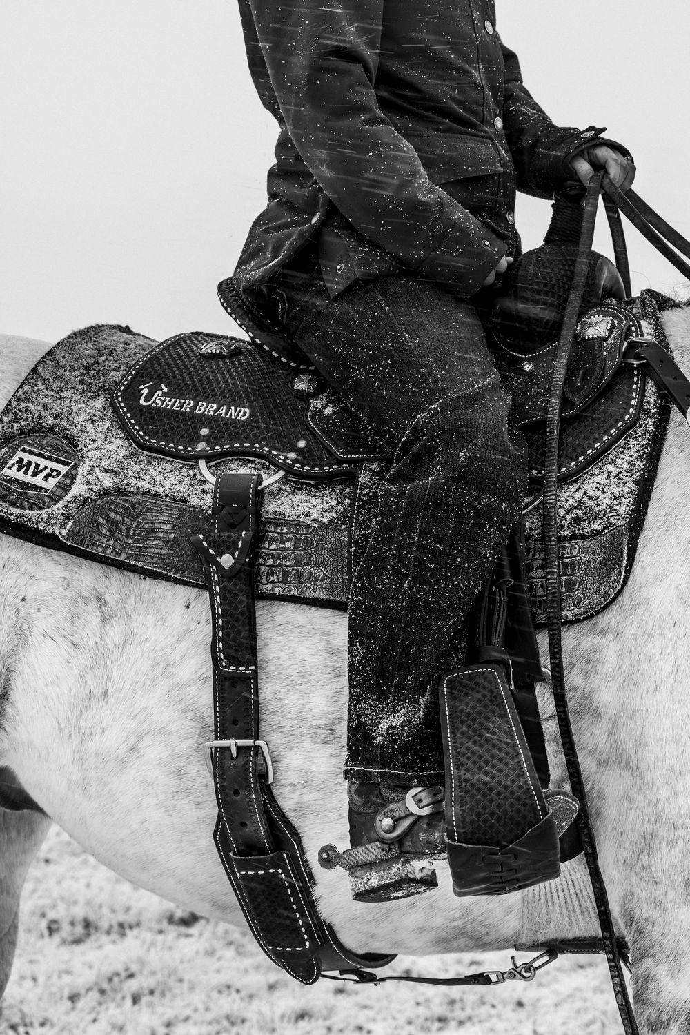 Black and white detail shot of a western saddle and rider's leg against a horse's flank.