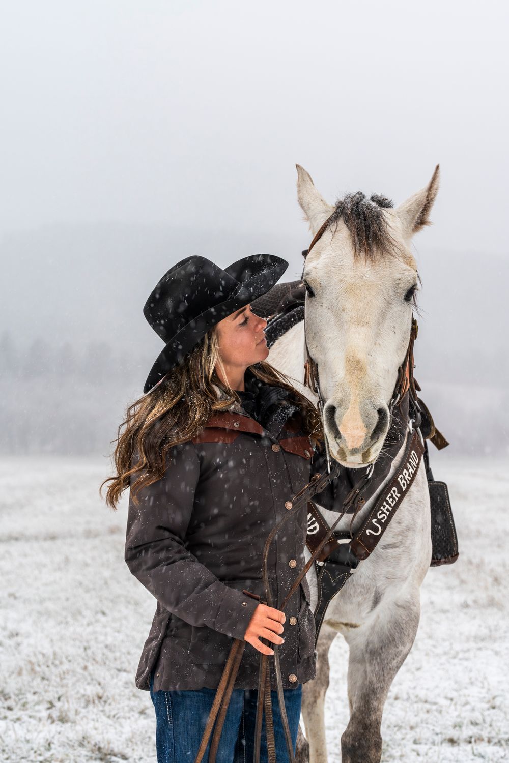 A cowgirl and her white horse share an affectionate moment during a snowy winter day.