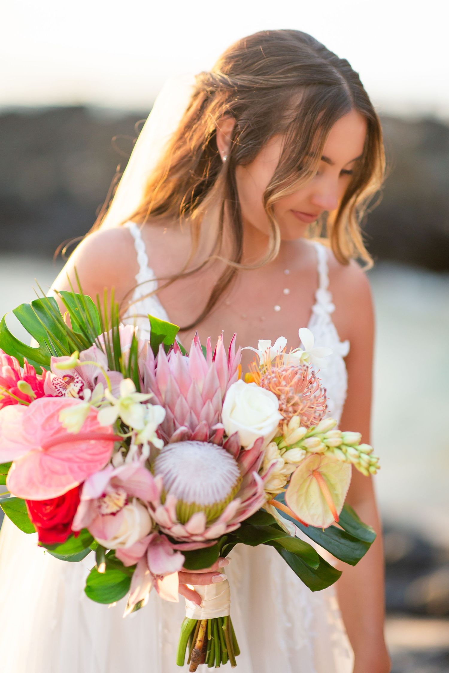 Bride in white lace gown holding tropical bouquet with pink protea and anthurium at beach sunset.