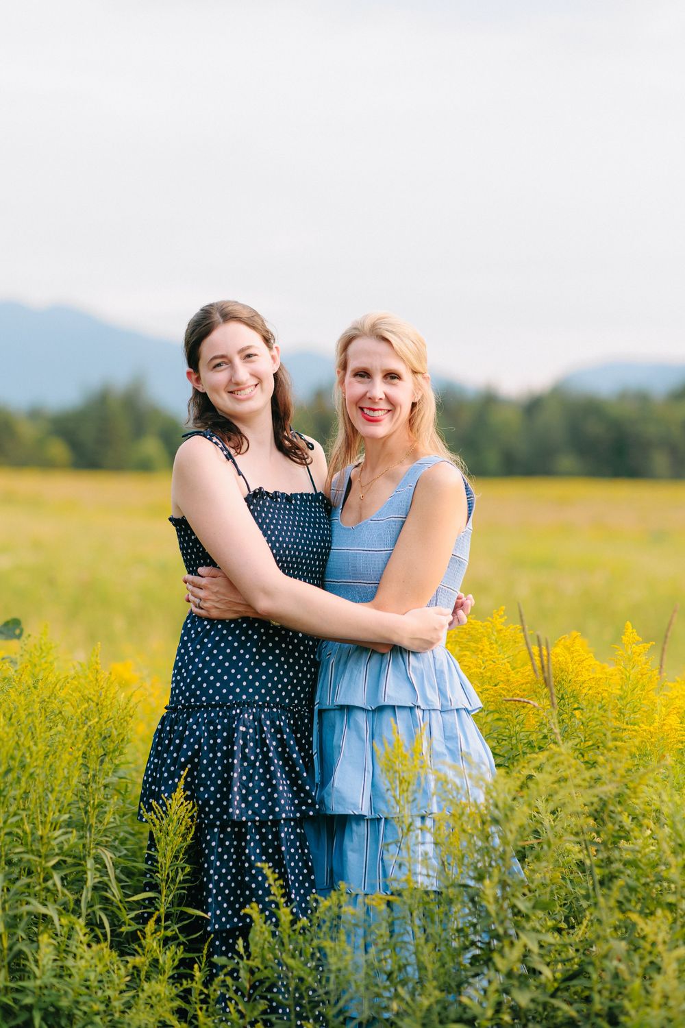 Two women in summer dresses embrace while standing in a blooming yellow wildflower field with mountains in the background.