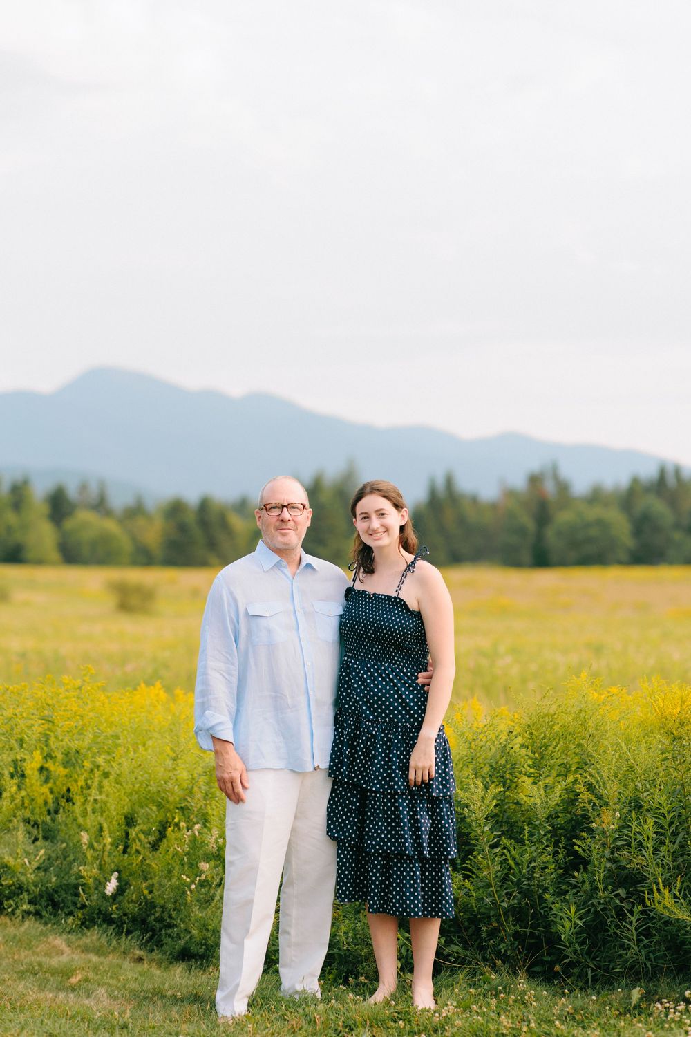 Two people pose together in a field of yellow wildflowers with mountains visible in the background.
