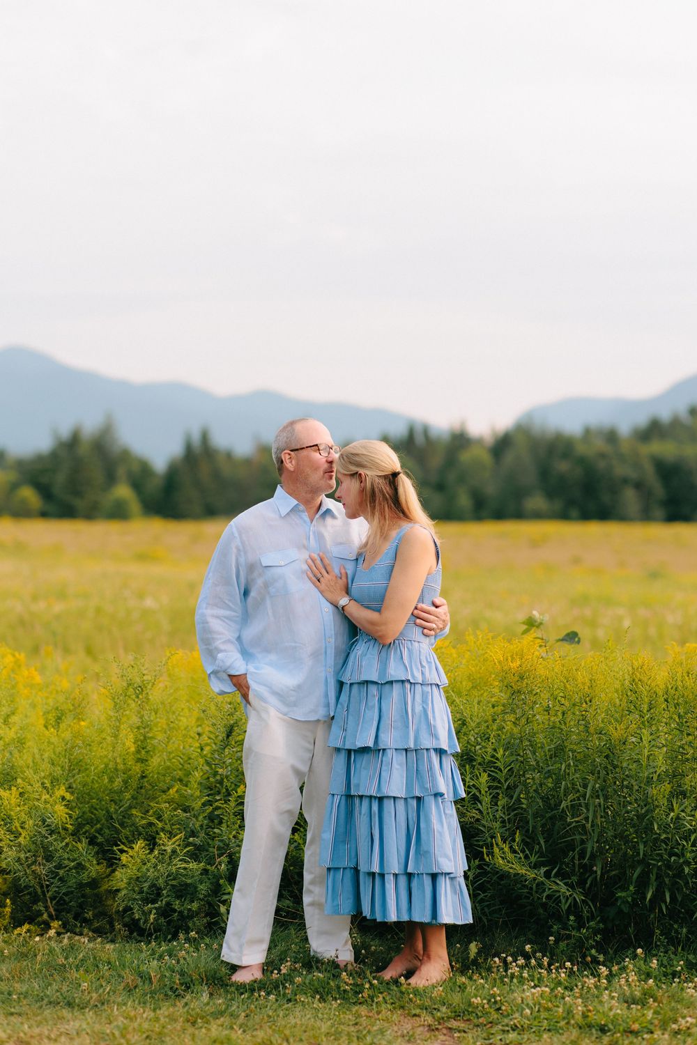 A romantic couple embraces in a field of yellow wildflowers with mountains and evergreen forest in the background.