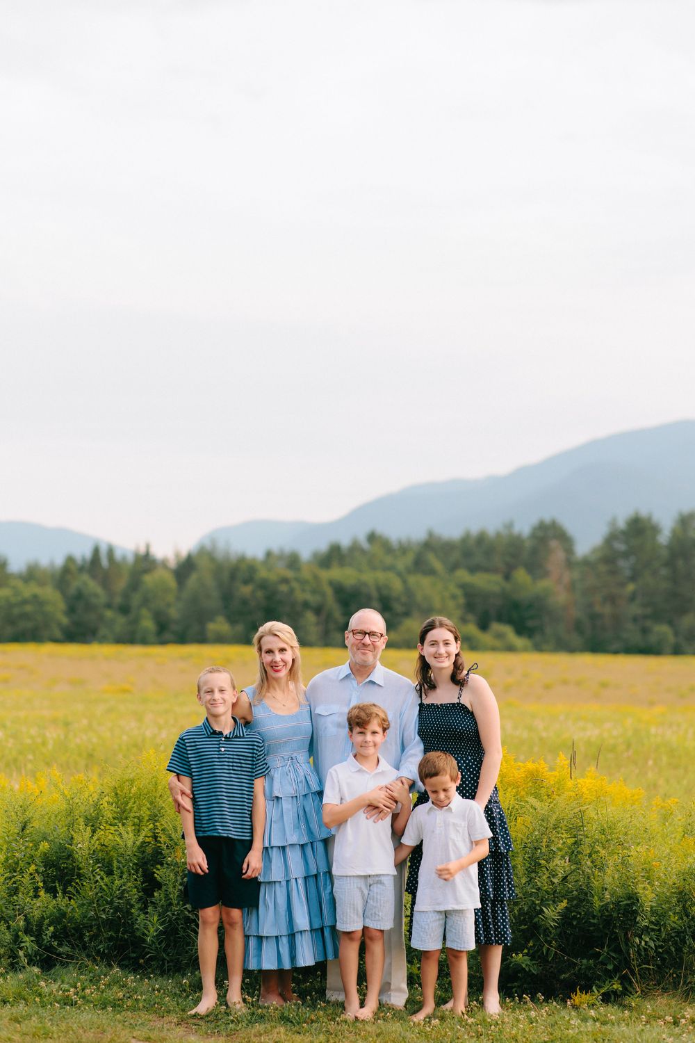A family stands together in a scenic mountain meadow with vibrant yellow wildflowers and evergreen forest in the background.