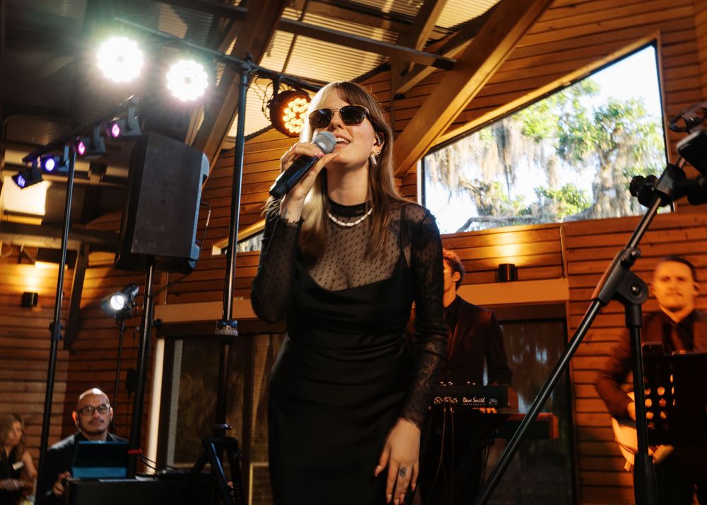 Female vocalist in black dress and sunglasses performs on stage at a wedding reception in a wooden venue.