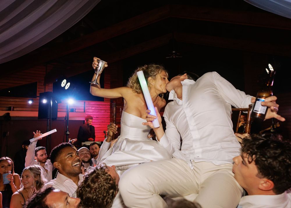 Bride and groom lifted by guests during lively wedding reception celebration with glow sticks and cheering crowd.
