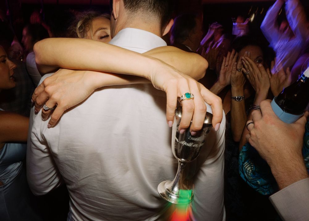 Couple embraces on crowded wedding reception dance floor, bride holding champagne glass with emerald ring visible.