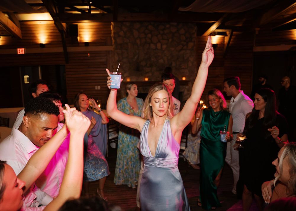 Guest in silver dress celebrates with arms raised on a crowded wedding reception dance floor.