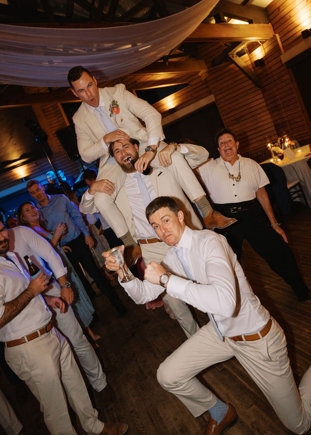 Groomsmen in tan suits pose playfully in a human pyramid on the wedding reception dance floor.