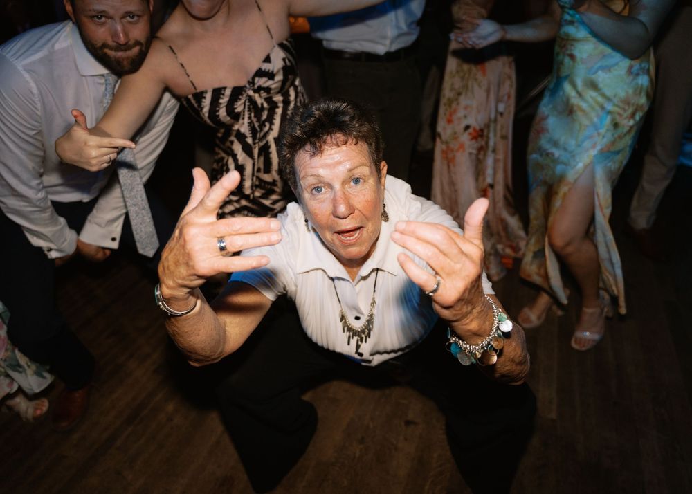 Energetic wedding guest dances enthusiastically on the reception dance floor, leaning toward the camera with hands raised.