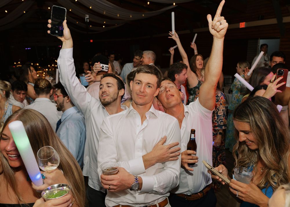 Excited wedding guests celebrate on the dance floor, raising hands and phones during lively reception party.