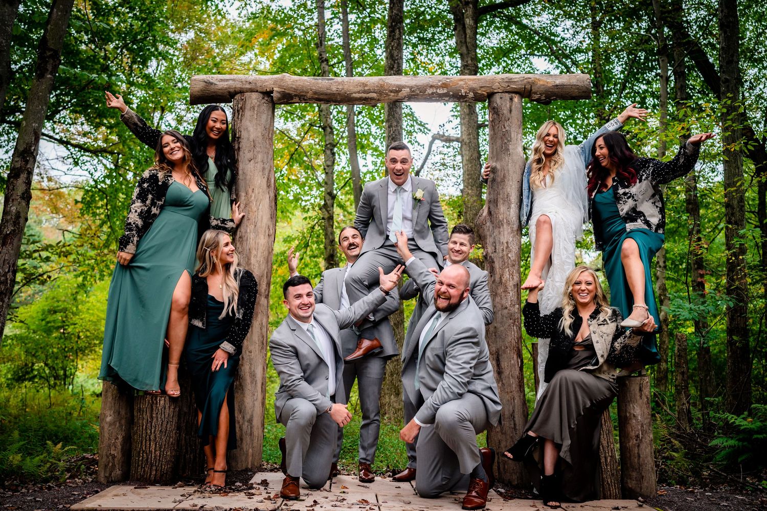 Wedding party poses playfully under a rustic wooden arch in a forest setting.