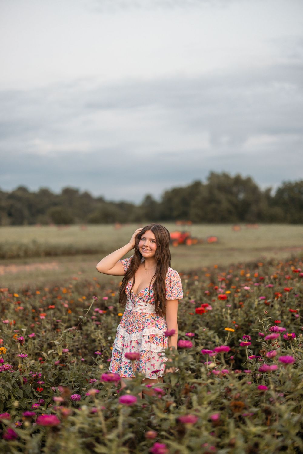 Person in floral dress stands among vibrant pink and red flowers in a field during sunset.