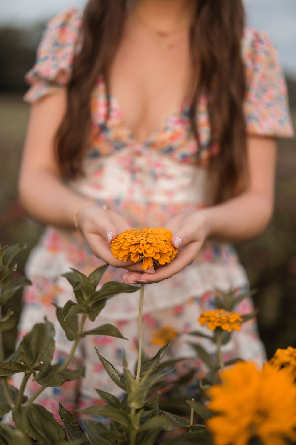 Person wearing a floral dress gently touches a vibrant orange marigold flower in a garden.