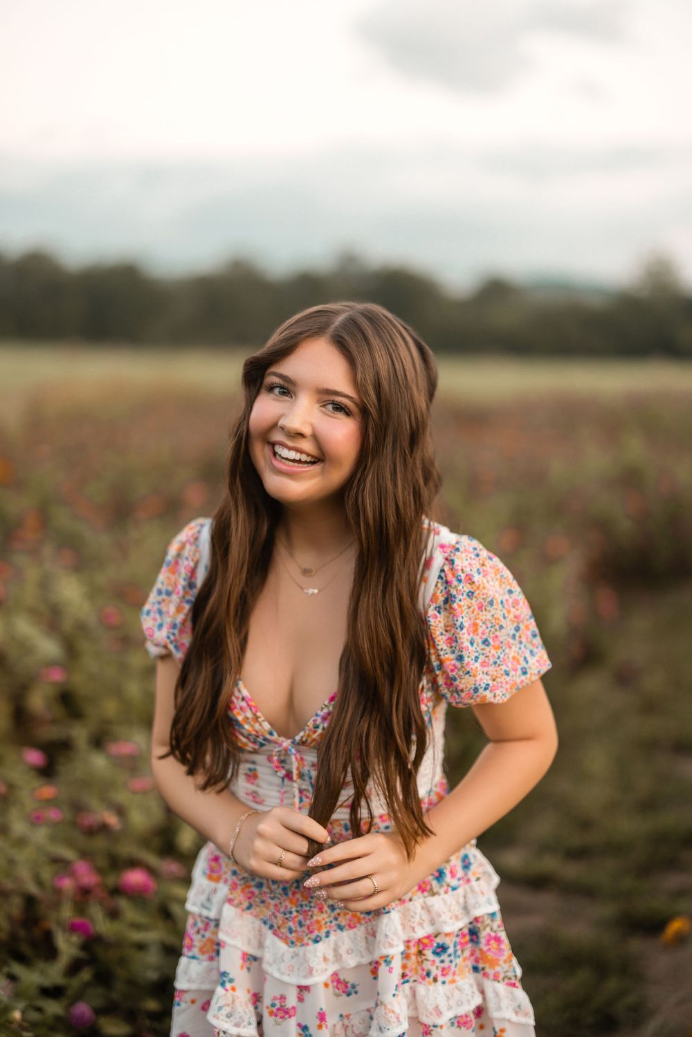 A smiling woman wearing a floral print dress poses in a flower field during golden hour.