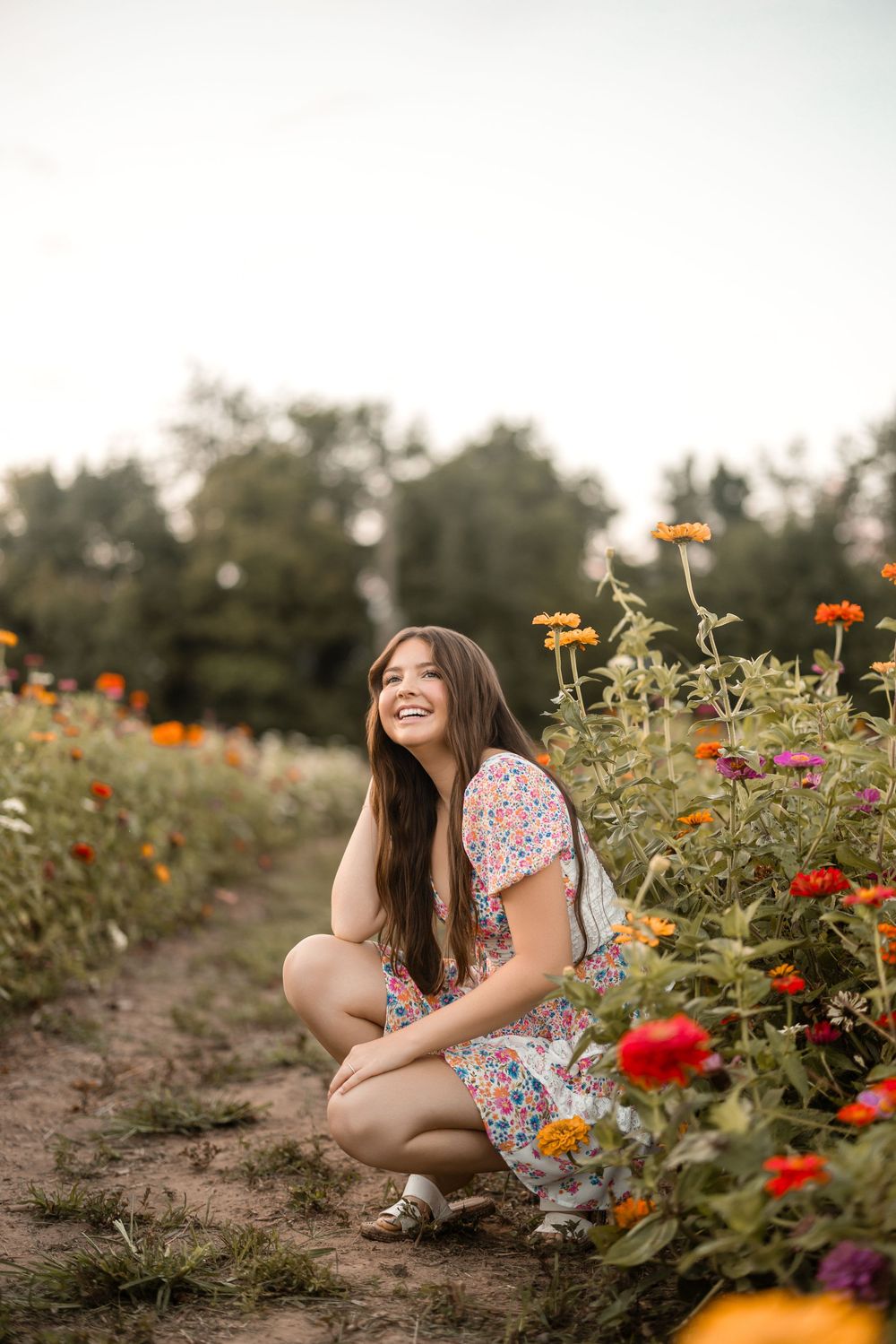 A woman in a floral dress sits among vibrant blooming flowers in a garden during golden hour.