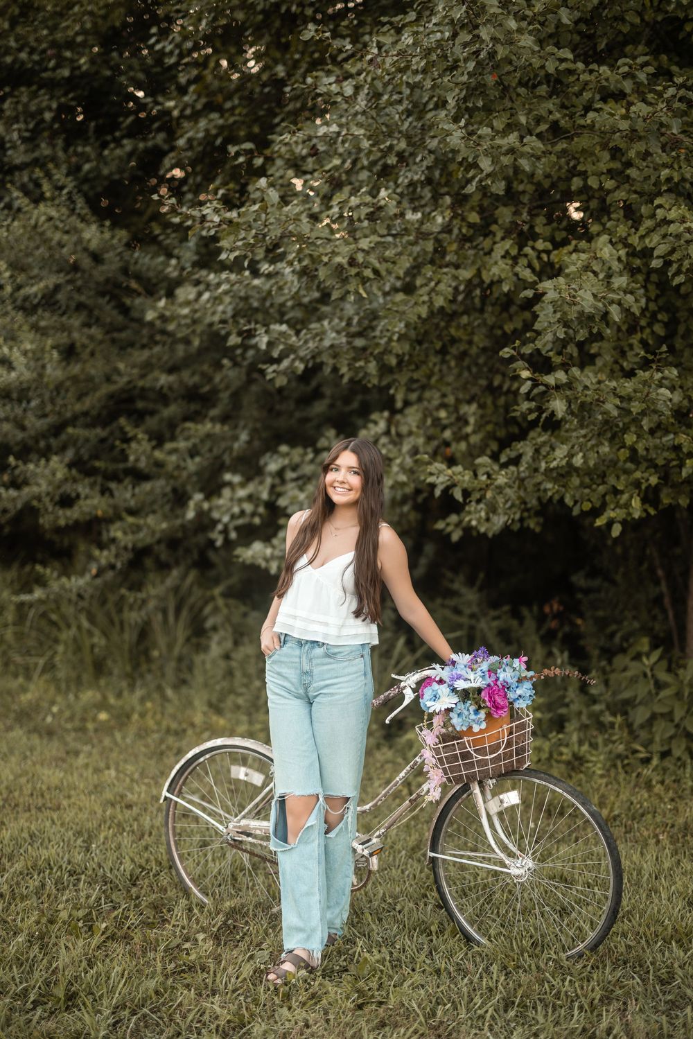 Vintage bicycle with flower basket stands beside someone in light summer attire against a lush green background.