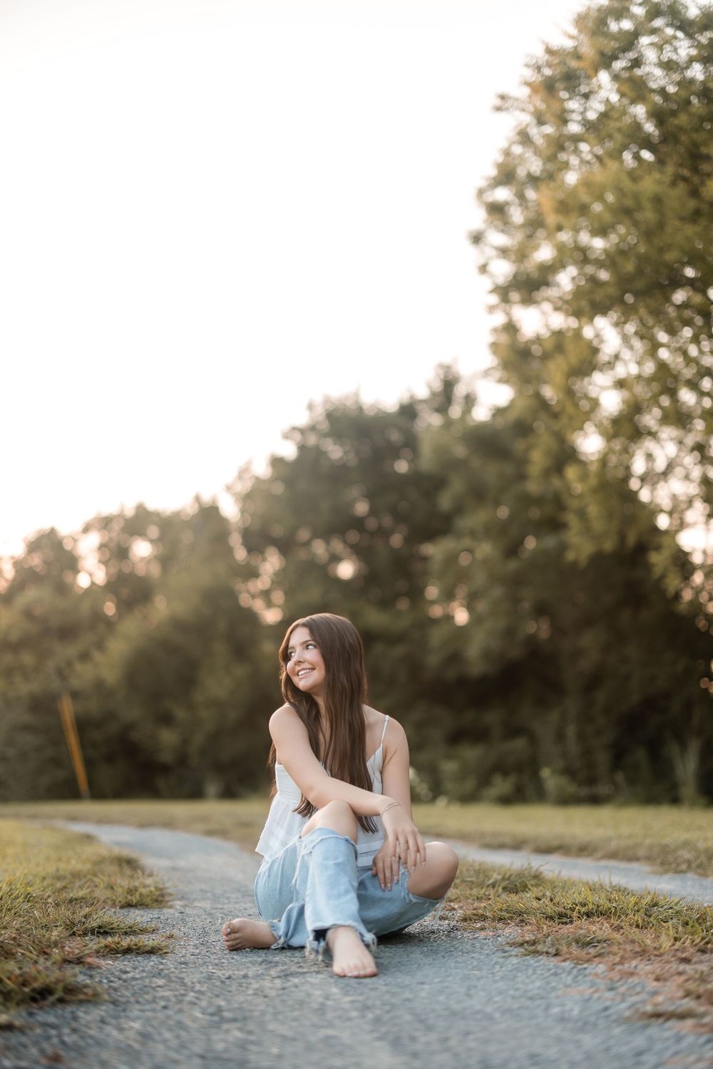 A person in a white top and light blue jeans sits casually on a dirt path during golden hour with trees in the background.