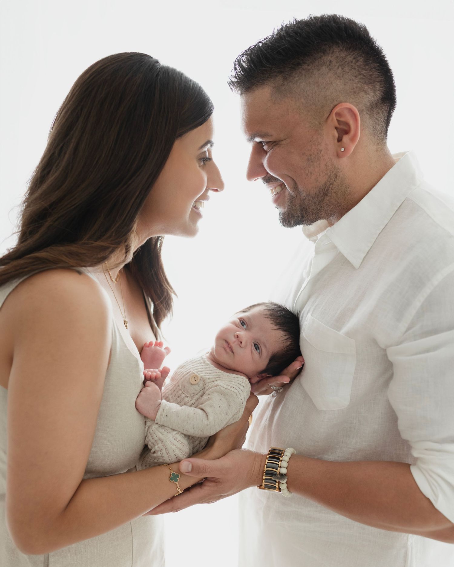 Couple in white outfits lovingly gazes at their newborn baby held between them against light background. Elizabeth Lang Photography.