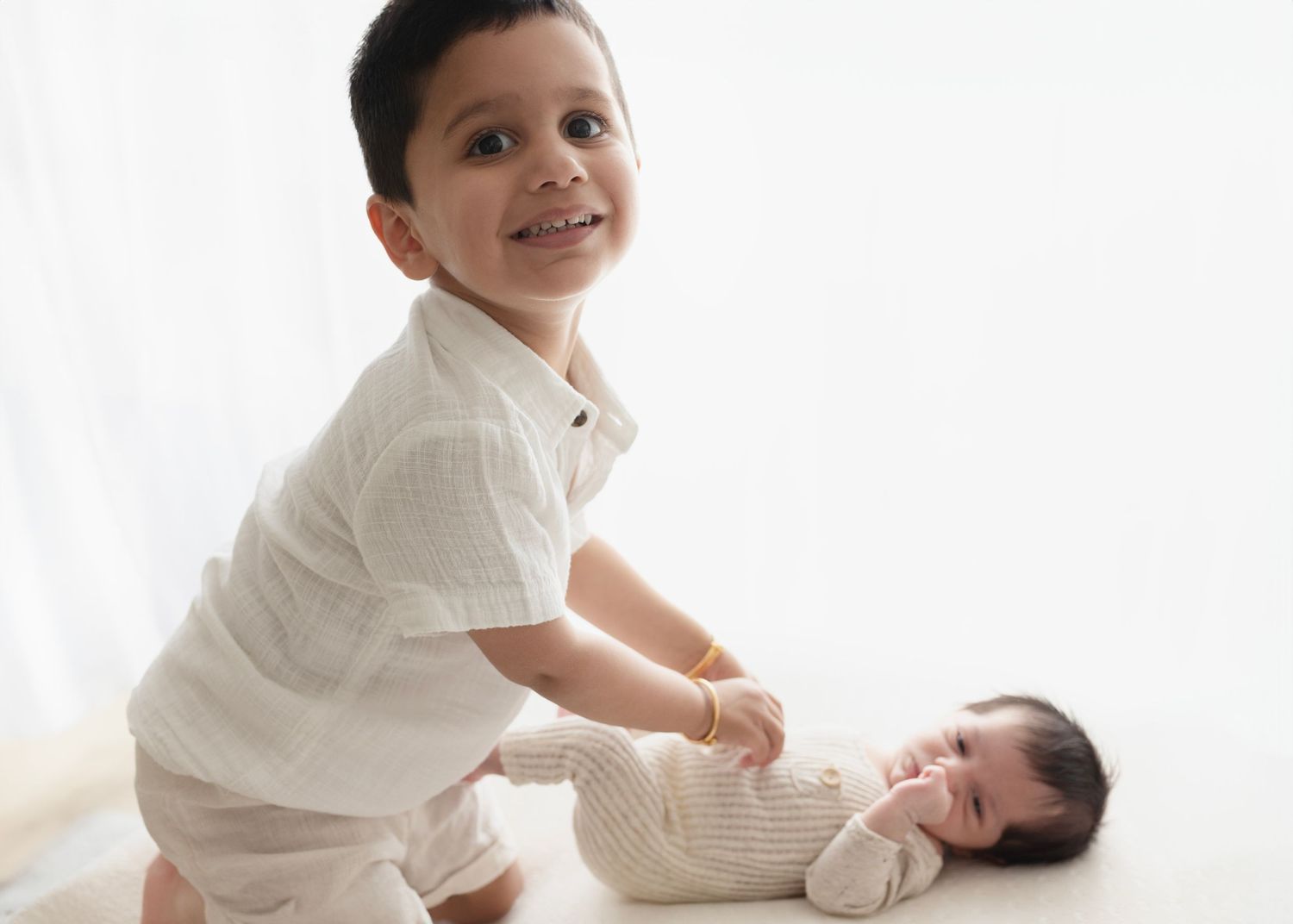 Two children on a white background, one sitting and one lying down, both wearing white clothing.