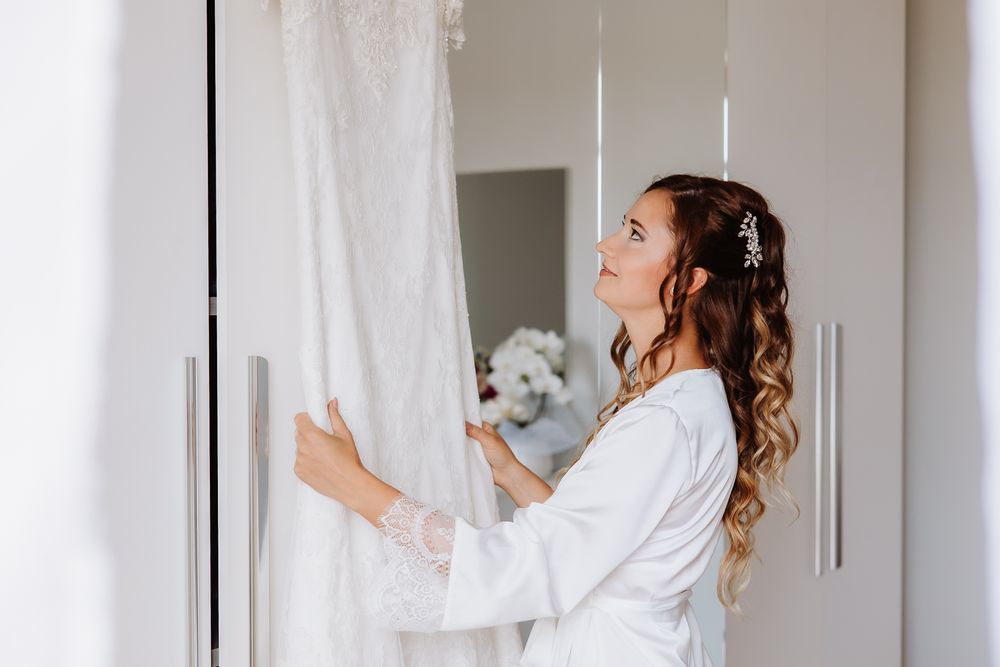 Person in white robe holding a small white dog while looking through curtains in a bright interior setting.