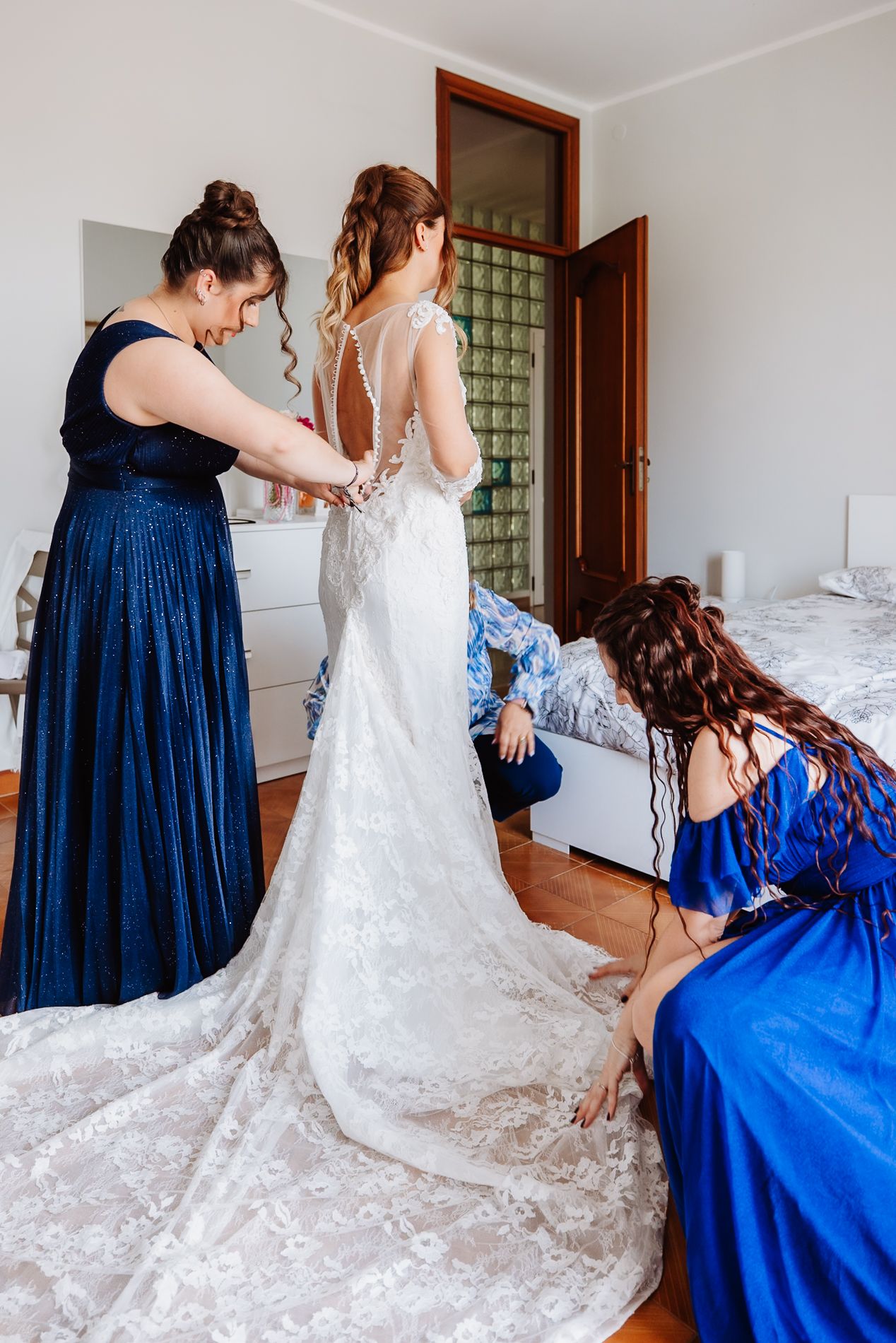 A bride getting ready in a stunning lace wedding gown with long train as attendants in blue dresses help with final details.