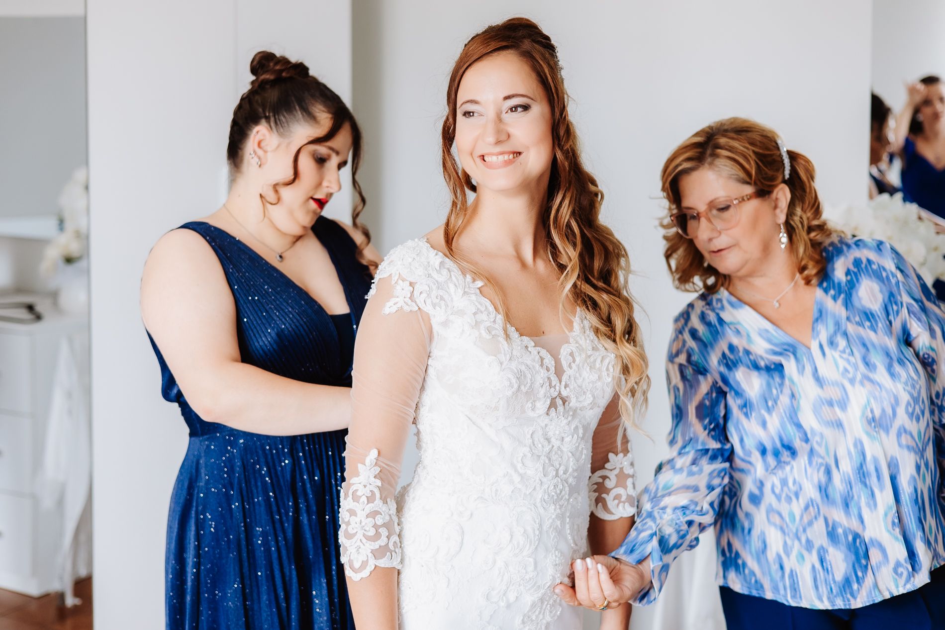 A bride wearing a long-sleeved lace wedding dress is assisted while getting ready with helpers in blue.