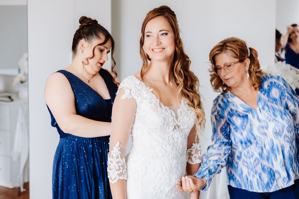 A bride wearing a long-sleeved lace wedding dress is assisted while getting ready with helpers in blue.