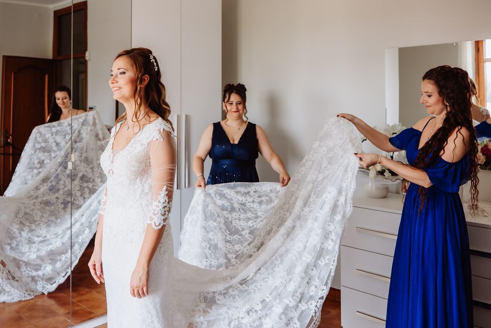 Bridesmaids in royal blue dresses help adjust a flowing white lace wedding veil in a bright room.