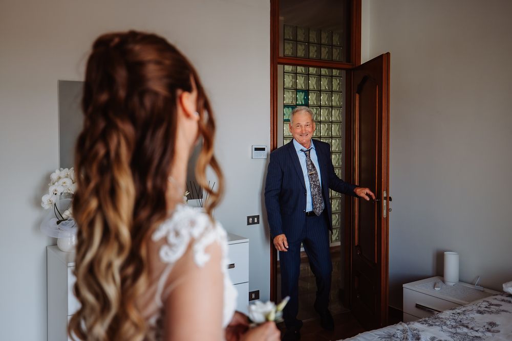 A bride with long wavy hair stands in a bedroom as someone enters through a doorway.