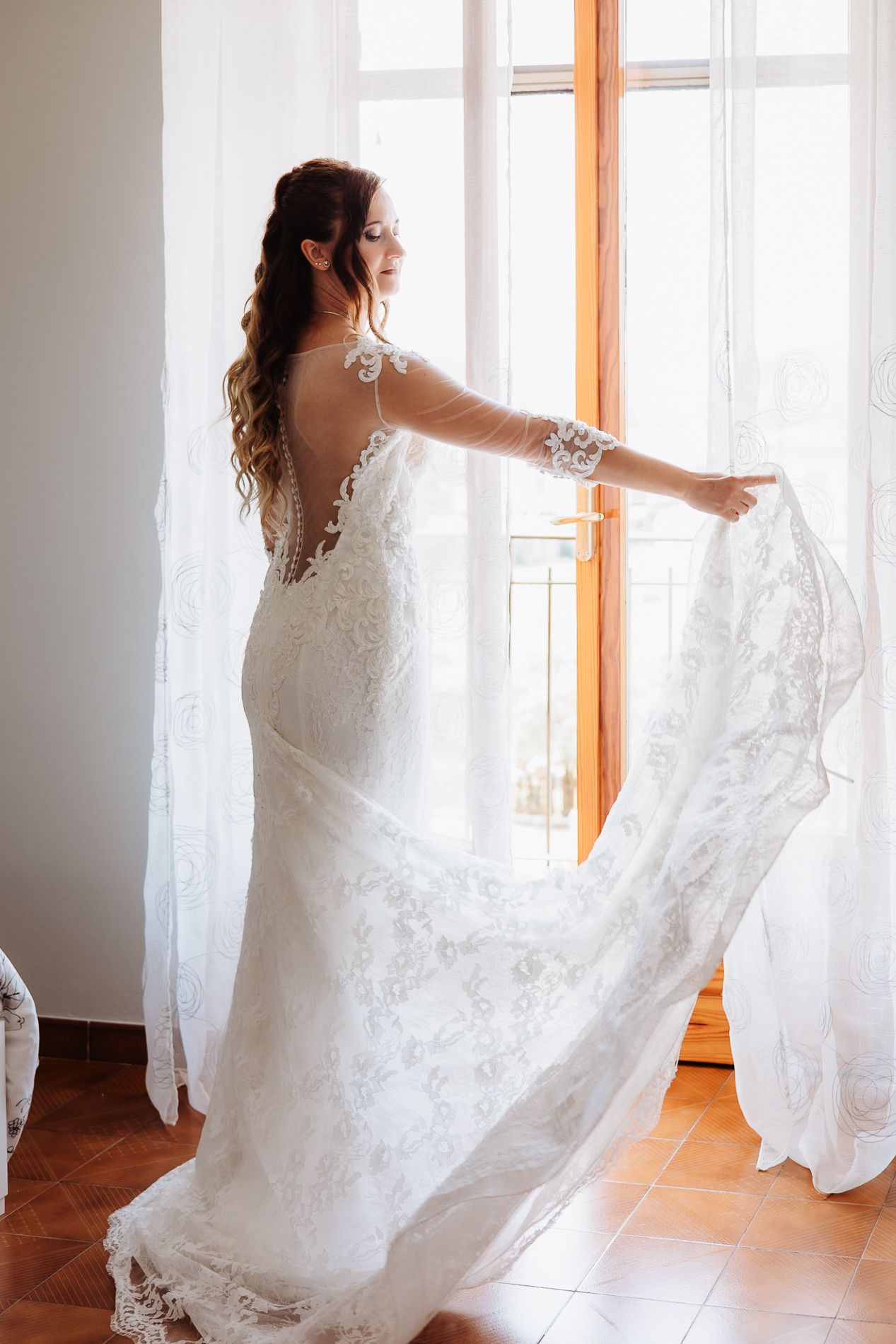 A bride in a flowing white lace dress twirls gracefully by sunlit windows, creating an ethereal bridal portrait.