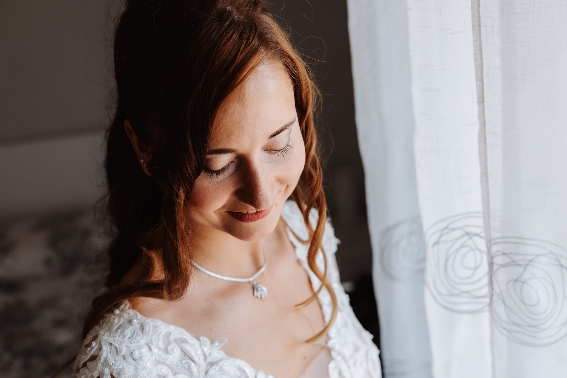Bride in a white lace gown with flowing hair stands next to sheer window curtains in natural light.