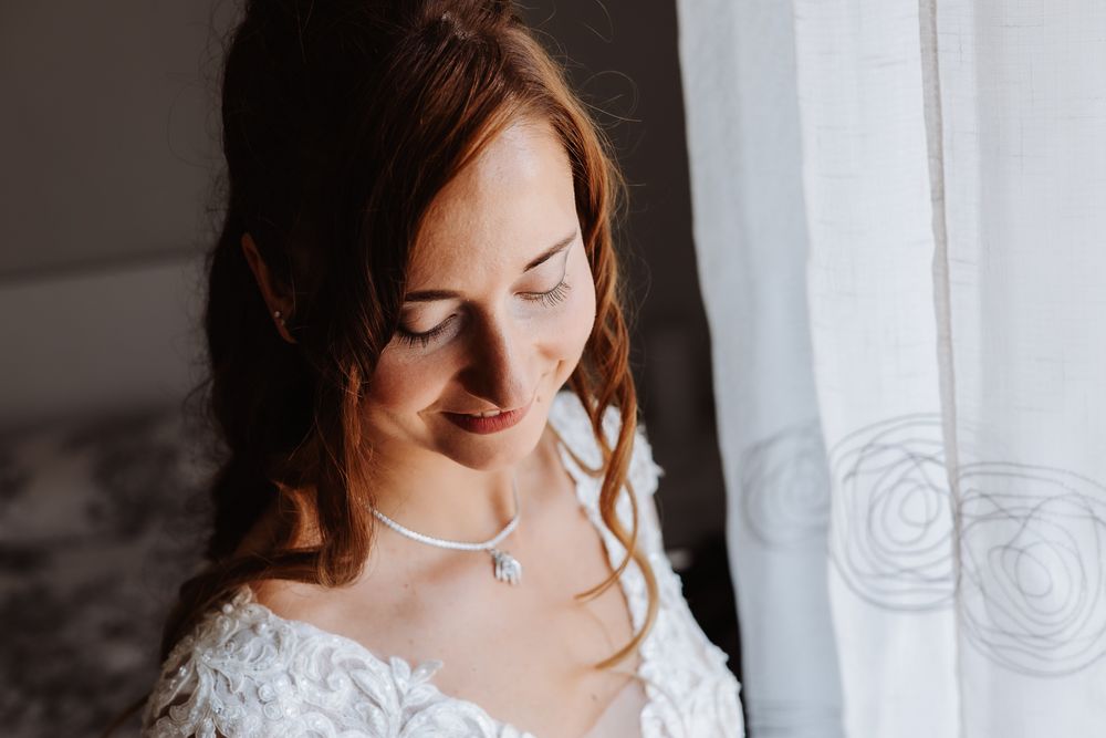 Bride in a white lace gown with flowing hair stands next to sheer window curtains in natural light.