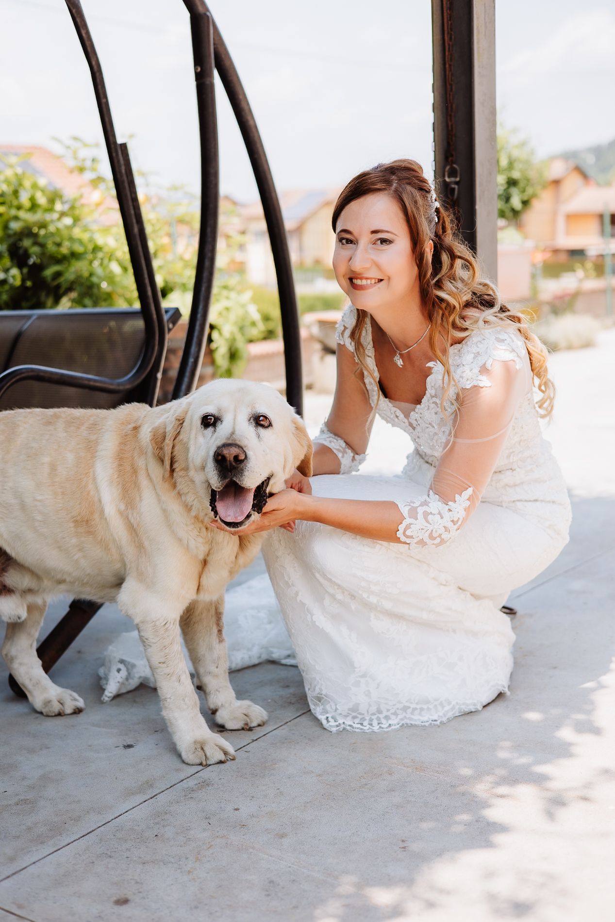 A happy yellow Labrador retriever poses with someone in a white lace wedding dress on a sunny outdoor porch.