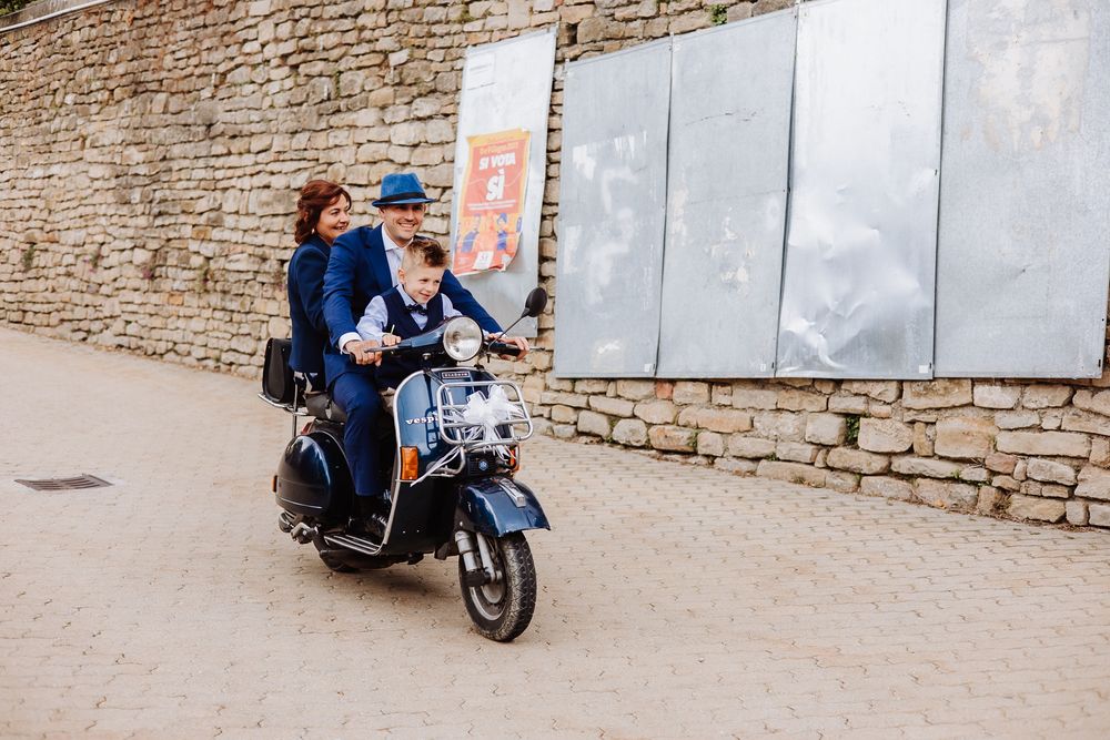 A vintage blue Vespa scooter traveling along a stone wall on a cobblestone street.