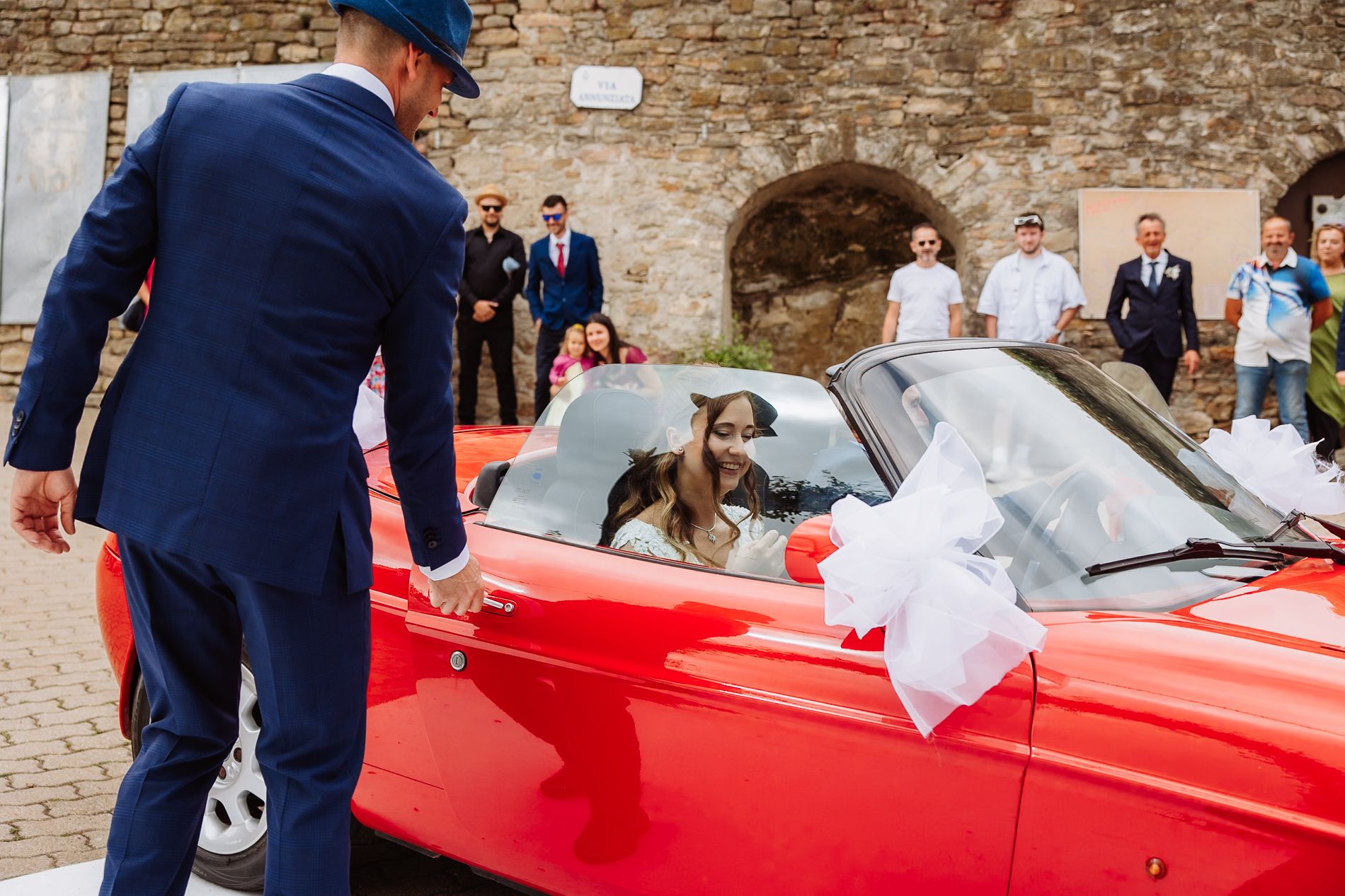 Wedding guests gather around a bright red classic Ferrari parked on cobblestone street during a celebration.