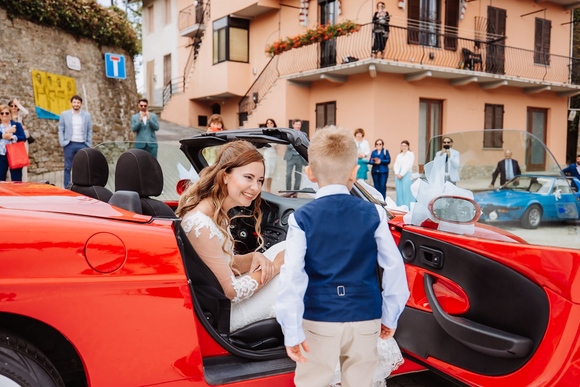 A bright red luxury sports car parked in a quaint European street with people interacting beside it.