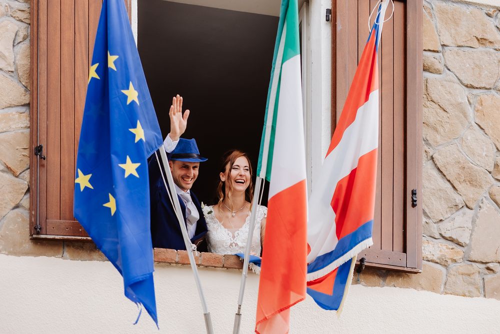 A couple stands together outdoors with wooden doors and European Union flags in the background.