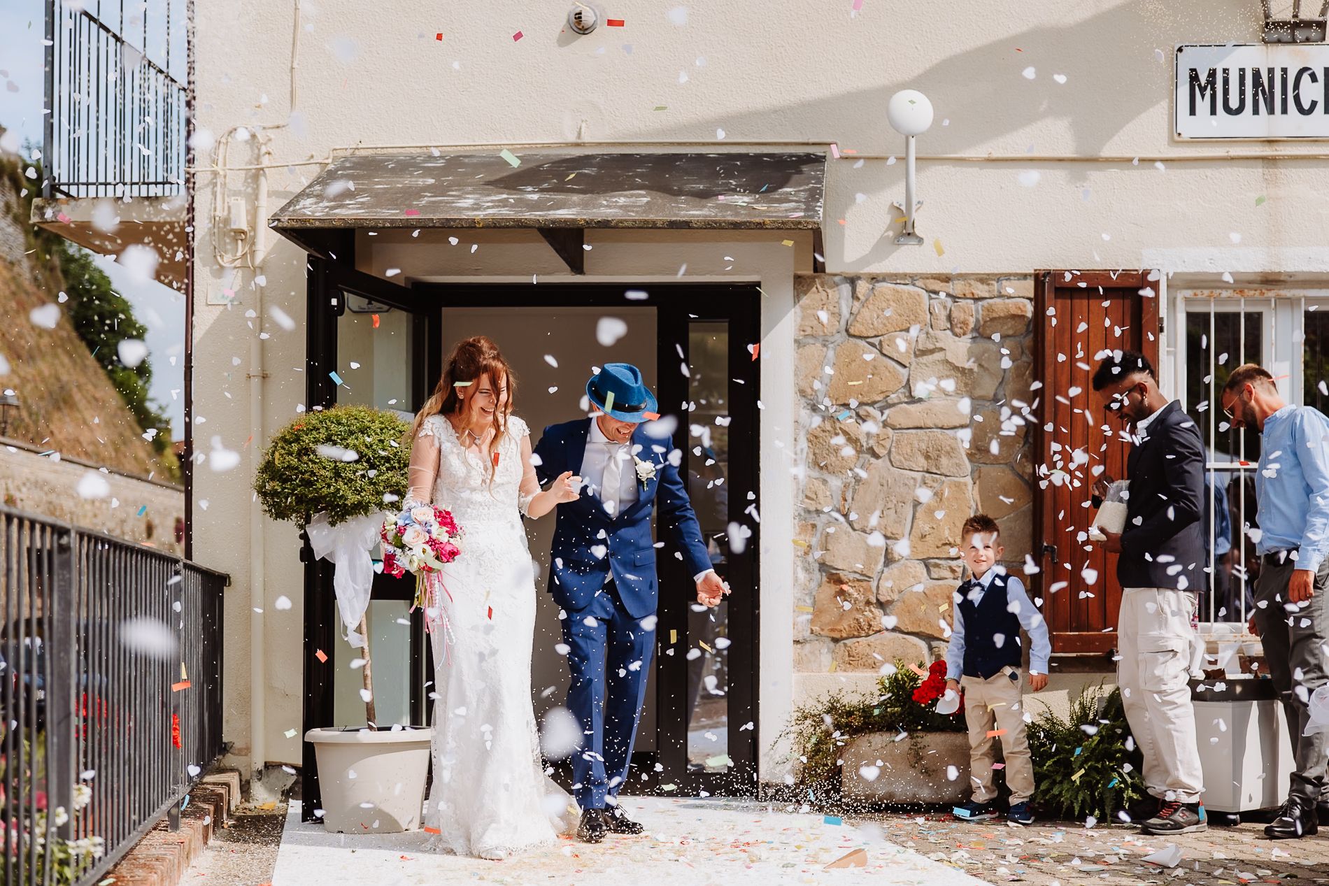 Wedding couple exits rustic stone church while guests throw flower petals in celebration during summer ceremony.