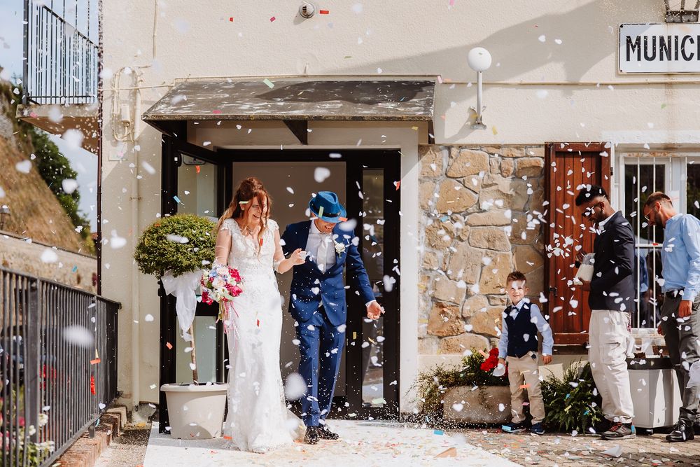 Wedding couple exits rustic stone church while guests throw flower petals in celebration during summer ceremony.