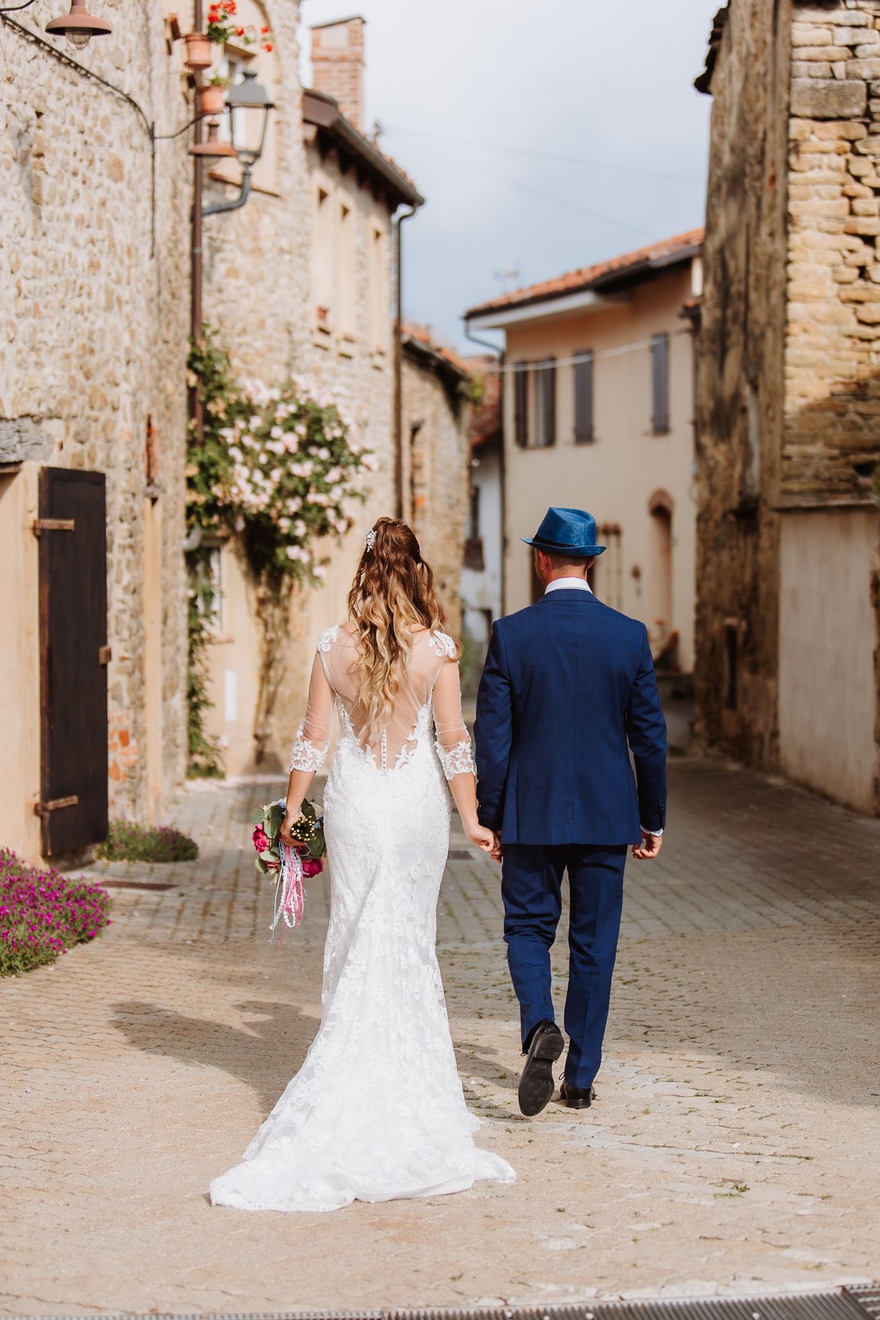 Newlywed couple walks hand in hand down narrow cobblestone street in charming European village with stone buildings.