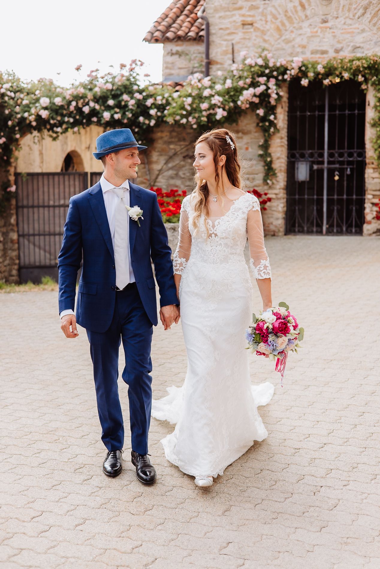 A newlywed couple walks hand in hand on a stone paved courtyard near a rustic stone building with climbing vines.