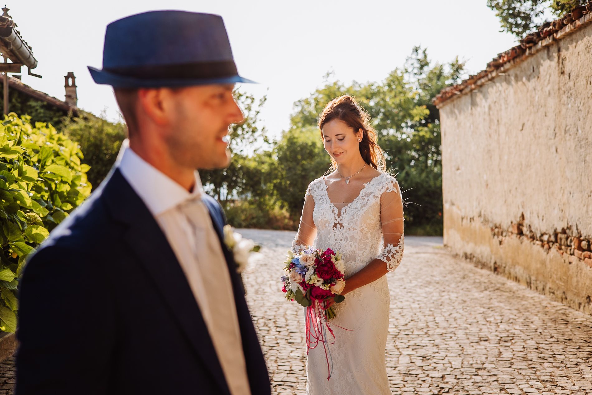 A wedding scene on a cobblestone path with a bride in white holding a bouquet and a groom wearing a fedora.