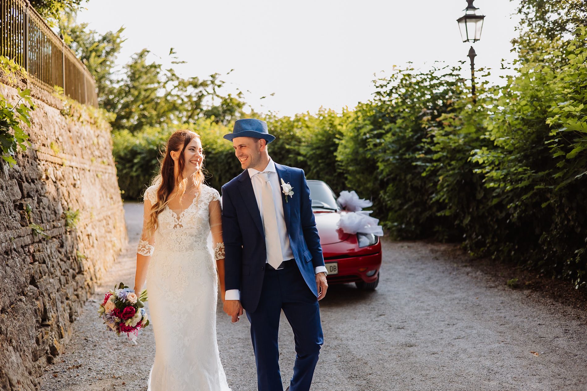 A couple walking together on a stone path with greenery on a sunny day while dressed in formal attire.