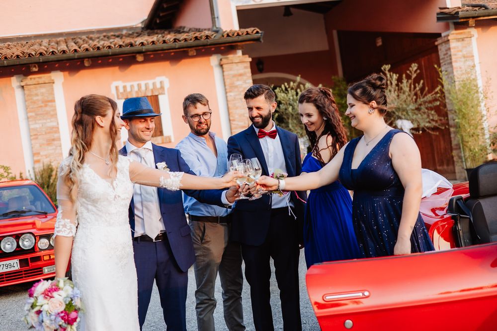 Wedding guests celebrate and take photos during an outdoor reception near brick buildings and a classic red car.